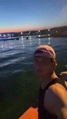 A selfie of a person wearing a cap and tank top standing outdoors near water at dusk, with a pier visible in the background.