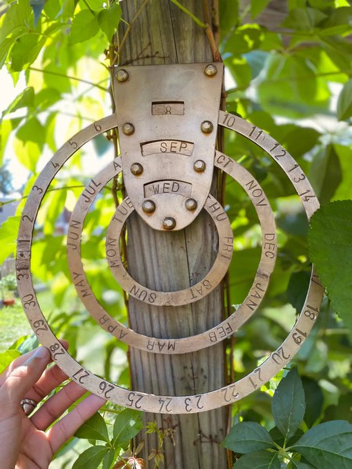 A hand holds up a decorative metal calendar ring set mounted on a wooden post, displaying months, days, and dates engraved in a spiral pattern.