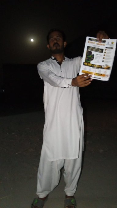 Muhammad Waqas, wearing a white traditional shalwar kameez, stands outdoors at night holding up a promotional flyer that reads "Earn more," showcasing his local outreach efforts for the AILYTIS and ODBIT community expansion in Kot Addu, Pakistan.