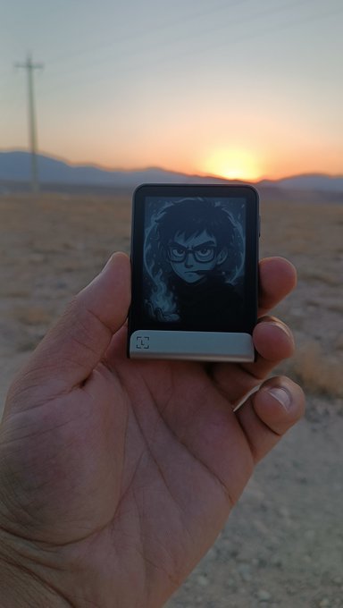 A hand holds a small device displaying an anime-style portrait of a bespectacled character, set against a desert landscape with mountains, a power line, and a setting sun in the background.