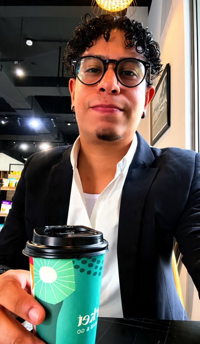 A man with curly hair, glasses, and a goatee smiles at the camera while wearing a navy blazer over a white shirt, holding a green paper cup labeled '19LAN' with a sunburst design in what appears to be a cafe.