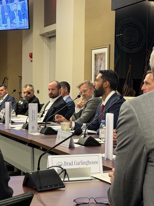 A panel of men in business attire sit at a long table during a formal meeting, with a visible nameplate reading "Brad Garlinghouse" and a monitor displaying the same speaker. The setting appears to be an official government or regulatory hearing, consistent with the post describing a speaking engagement at the inaugural CFTC Innovation Advisory Committee meeting.