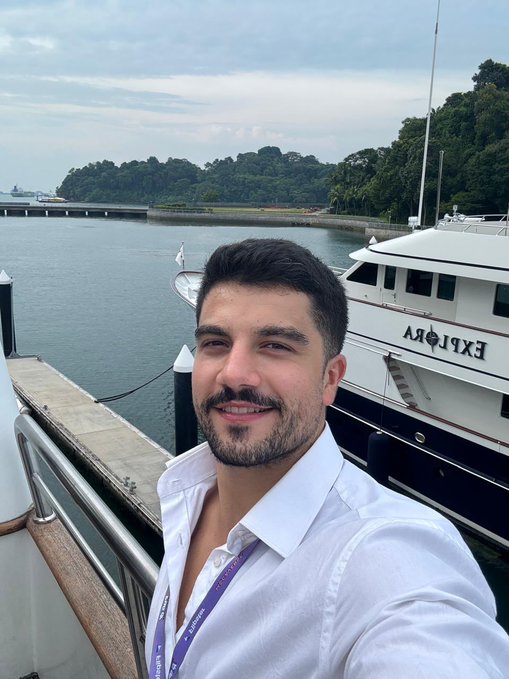 A selfie of a man in a white shirt on a dock, with a yacht named 'EXPLORA' and a forested coastline in the background.