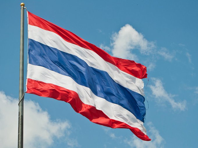 The flag of Thailand, featuring red, white, and blue horizontal stripes, waves against a partly cloudy blue sky.