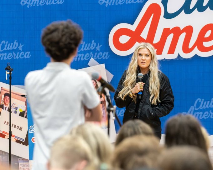 A blonde woman in a black jacket speaks into a microphone on stage at a 'Club America' event with a blue backdrop, while a young boy holds up papers in the foreground and an audience watches.