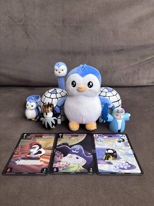 A large Piplup Pokémon plush sits on a gray cushion surrounded by smaller Pokémon figures, with three trading cards displayed in front of them.
