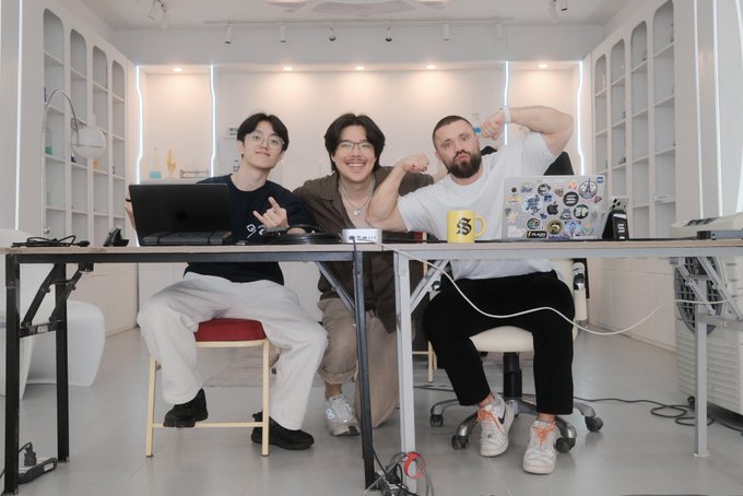 The image shows three people sitting at a table, each with their own laptop open in front of them. They are all smiling and appear to be enjoying themselves while working or socializing together. The room they're in has white walls and a gray floor, giving it a clean and modern look. There is also a chair visible near the table where the people are seated.