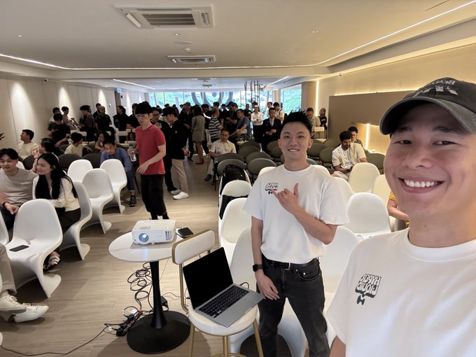 Two smiling young men wearing white t-shirts and caps pose for a selfie in a bright, crowded event space with rows of white chairs, a projector on a small table, and a laptop.