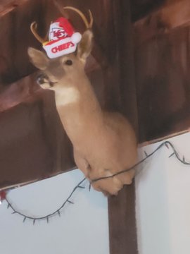 A decorative deer figure wearing a small Santa hat and reindeer antlers sits perched on a wooden beam.