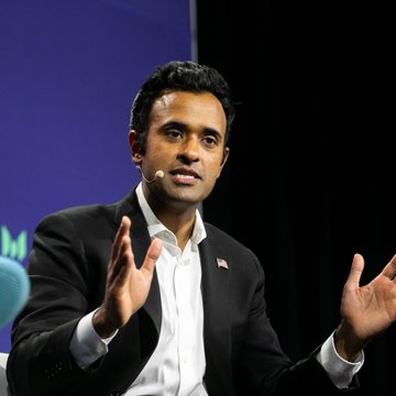 A man in a dark suit speaking at a panel or conference, gesturing with both hands while wearing a lapel microphone.
