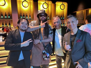 Four men pose together in a warmly lit venue with circular wall decorations, one wearing a hat and sunglasses in the center.