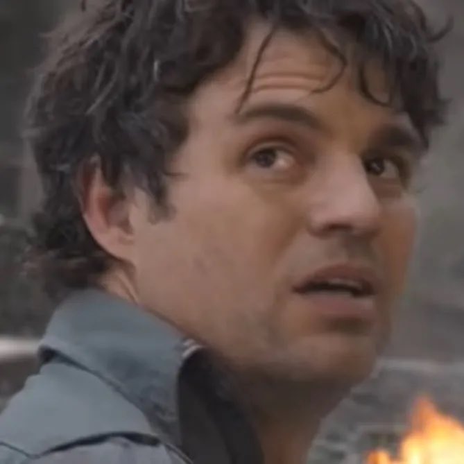 A close-up reaction image of a curly-haired man looking surprised or shocked, with flames visible in the background.