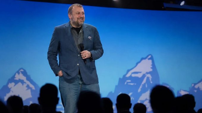 Charles Hoskinson, founder of Cardano, holds it on stage during a keynote presentation, smiling while speaking into a microphone, likely discussing the upcoming "Midnight" network and agentic trading features.
