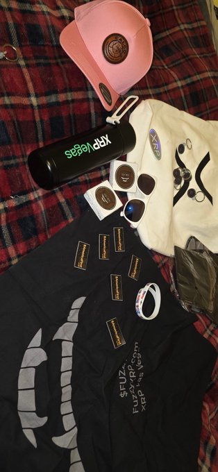 A black t-shirt and cream-colored beanie, both branded with "FUZZY x XRP" text, are displayed alongside a pink Fuzzy cap, an XRP-branded water bottle, sunglasses, pins, wristbands, and other giveaway items received as a one-year anniversary gift from Fuzzy at XRP Las Vegas.