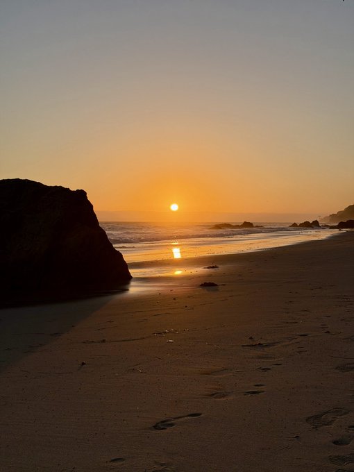 A serene sunset scene at a beach, with the sun low on the horizon casting a golden reflection on the water, framed by a large rock silhouette on the left and footprints in the sand in the foreground.