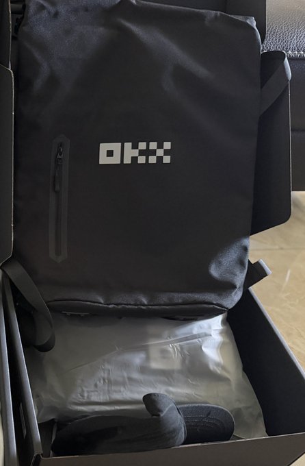 A photo of an unboxed black OKX branded backpack sitting inside its shipping box, with packaging material and a glove visible at the bottom.