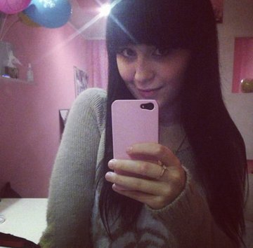 A young woman with long dark hair takes a mirror selfie using a pink phone in a pink room.
