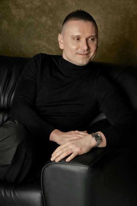 A man wearing a black turtleneck sits on a black leather couch with his hands folded, wearing a wristwatch.