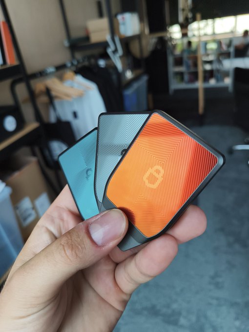 The image shows three electronic devices being held up by a person's hand against a gray background, with a box visible behind the devices. The devices are square-shaped and have different colors - one orange, one blue, and one black. The person is holding them in their hand, possibly considering which device to purchase or displaying them for others to see.