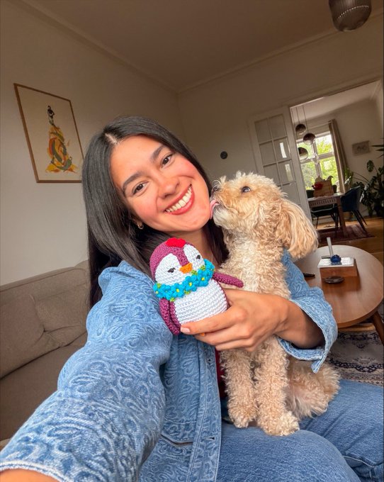 A woman taking a selfie while holding a small dog that is licking a crocheted penguin plush toy, in what appears to be a living room setting.