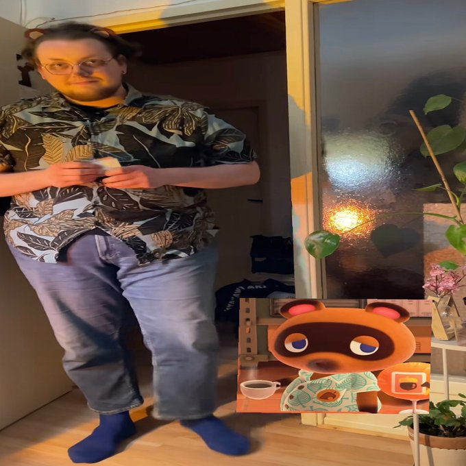 A man wearing glasses, a patterned shirt, and jeans stands indoors holding a mug, with an inset image of the video game character Tom Nook from Animal Crossing.