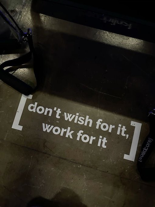 A motivational quote "[ don't wish for it, work for it ]" with "solidcore" branding painted on a gym floor, viewed in dim lighting near some workout equipment.
