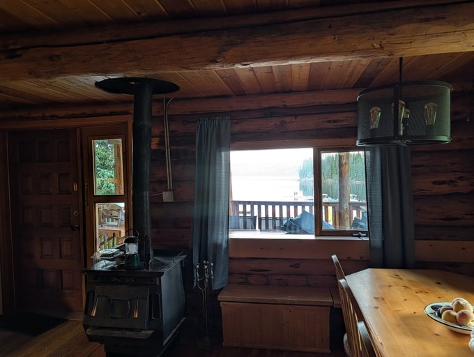 Interior of a rustic wooden cabin featuring a black wood-burning stove, a window with a view of water, and a dining table.