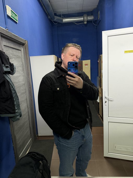 A man taking a mirror selfie in a hallway with blue walls, wearing a black jacket and jeans, holding a phone with a blue/red case.