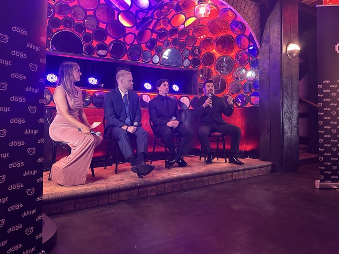 Four people sit on a stage at an event branded 'dogs of dwnyc' (visible on side panels), with a decorative wall of circular plates illuminated by pink and purple lighting behind them.