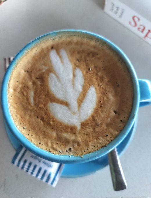 A blue coffee cup featuring a beautifully crafted leaf latte art design sits on a saucer, with a spoon and sugar packets nearby on a table. The image reflects the caption's theme of taking a brief, peaceful moment to breathe and recharge before tackling a "Variational points drop" in the morning.