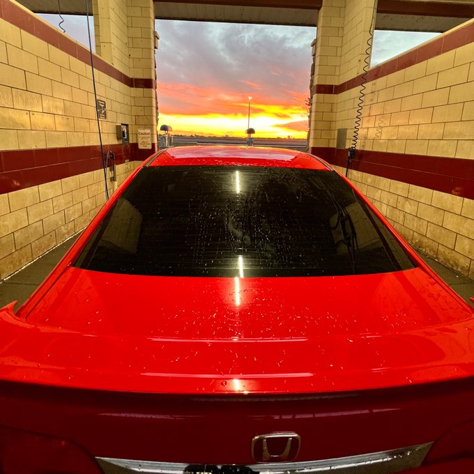 The rear of a red Honda car is parked in a car wash bay, with a vibrant orange and pink sunset visible through the opening.