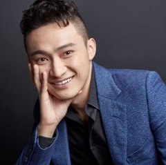 Studio portrait of a smiling young Asian man with short dark hair, wearing a dark shirt under a blue blazer, resting his chin on his hand against a dark gray background.