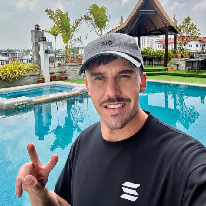 The image features a man wearing a black t-shirt with an "EOS" logo, standing by a swimming pool. He is making a peace sign gesture while posing for the photo. The background of the image shows a beautiful view of palm trees and buildings, suggesting that he might be in a tropical location like the Philippines.