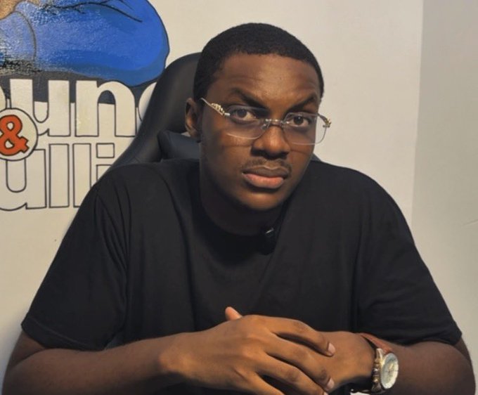 A young man wearing glasses and a black t-shirt sits with his hands clasped in front of a wall with partially visible text 'un...' and '...ui...'.