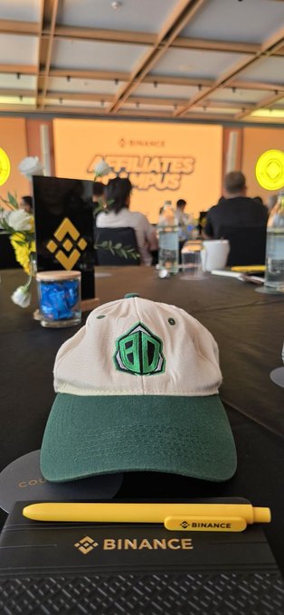 A cream and green baseball cap with a "BC" logo sits on a Binance-branded notepad alongside a yellow Binance pen, with the "Affiliate Campus" stage and Binance signage visible in the background—capturing the vibrant atmosphere of Day 2 at Binance Campus Asia 2026.