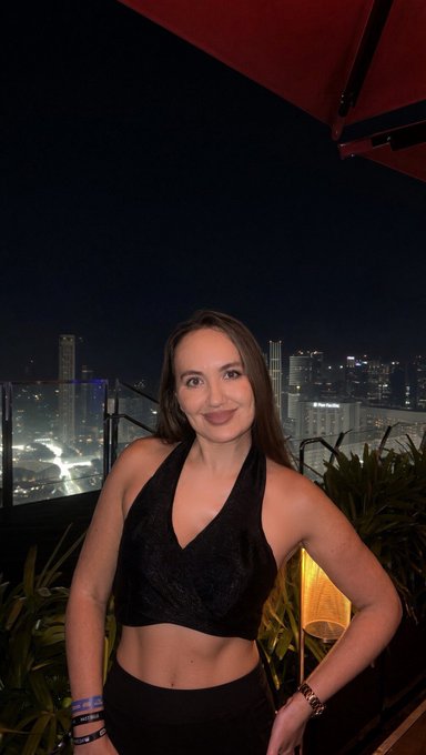 A woman with long brown hair poses at night on a rooftop or balcony overlooking a city skyline, wearing a black halter crop top and a watch on her wrist.