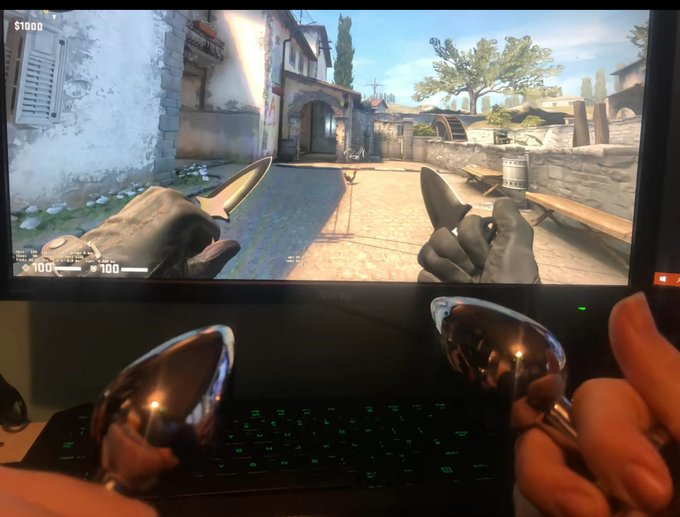 A first-person photo of someone holding two metal spoons in front of a laptop displaying a first-person shooter game, creating an improvised controller setup.