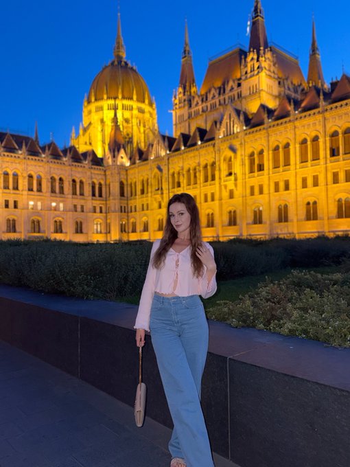 A young woman in a pink blouse and jeans poses in front of the illuminated Hungarian Parliament Building in Budapest at dusk.