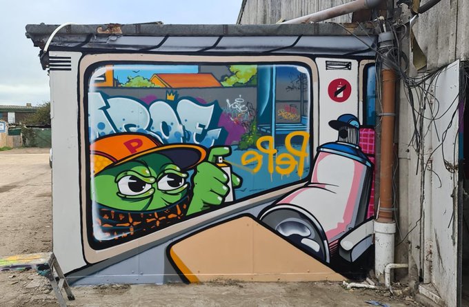 A colorful street art mural painted on the side of a building, depicting a green cartoon character wearing an orange cap with a "P" holding a spray paint can, alongside graffiti text.