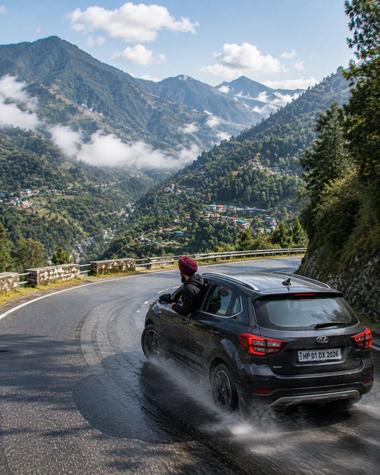 A photograph of a person leaning out of the window of a dark grey Hyundai car driving on a winding mountain road, with a scenic view of a lush green valley and a town below, and a license plate reading "HP 01 DX 2026."
