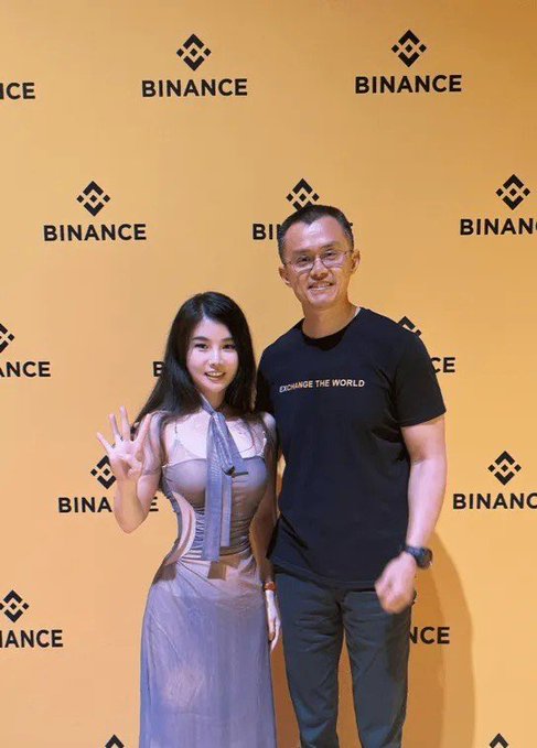 Two people pose in front of a yellow backdrop covered in Binance logos; the man wears a black t-shirt with the text 'EXCHANGE THE WORLD.'