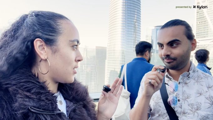 Two people having a conversation outdoors with modern glass skyscrapers in the background; the woman has curly hair and hoop earrings, while the man wears a patterned shirt and holds sunglasses.