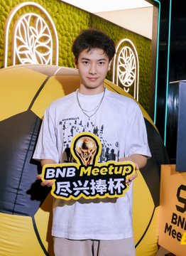 A young man stands in front of a yellow display with 'BNB Meetup' text, wearing a graphic t-shirt.