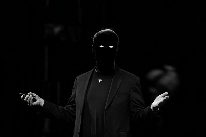 A black and white image of a person in a dark suit with glowing white eyes standing in shadow, holding their hands up while gripping a small object.