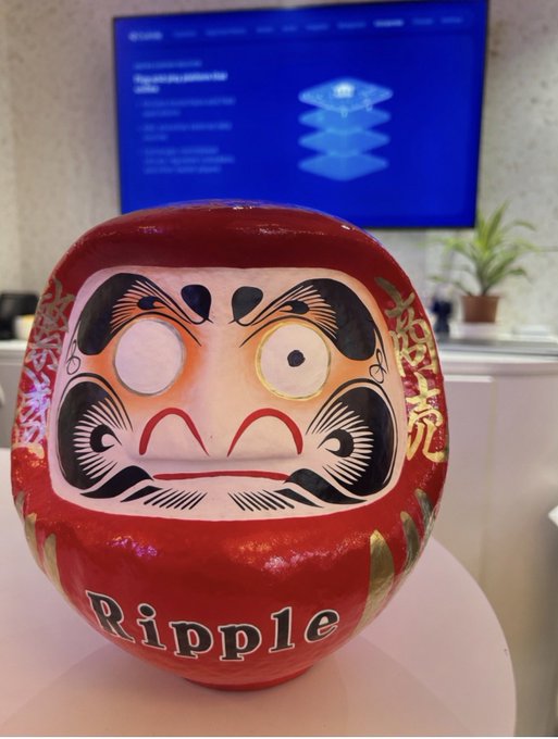 A red Daruma doll with the word "Ripple" painted on its base sits in front of a monitor displaying Ripple's website, embodying the post's message of resilience and hope for XRP's future with the Japanese proverb "Fall seven times, stand up eight times."