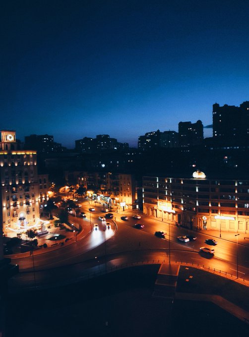 A twilight cityscape showing an illuminated roundabout with streaks of car headlights, framed by buildings lit against a deep blue evening sky.