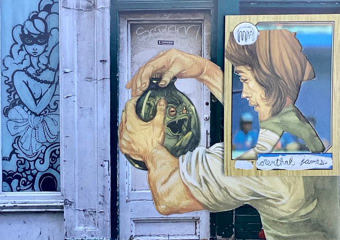 A street mural depicting hands squeezing a green blob with a screaming face, placed next to a framed portrait of a person looking down with the text "identical games" and "MOPSY" visible.