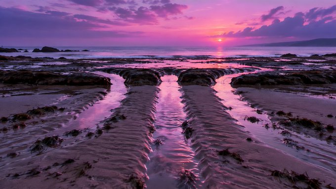 A stunning beach landscape at sunset with vibrant purple and pink skies, the sun setting over the ocean, and rippled sand channels reflecting the colorful light.