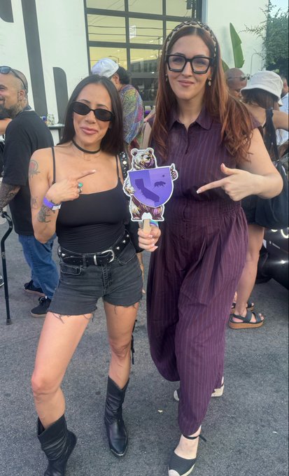 A photograph of two women posing together at an outdoor event; the woman on the right wears a purple jumpsuit and glasses, while the one on the left wears a black tank top, shorts, and boots, holding a sign with a bear graphic.