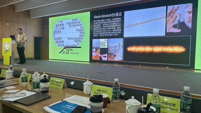 A presenter stands on stage at the 2026 International Symposium on Plastic Arts and Technological Media in Education held July 3 in Guangzhou, China, with slides displaying Kevin Abosch's work including "I AM A COIN (2018)" featuring cryptographic code and a coin image. The foreground shows a panelist table with name placards for Bernhard, Ryota, and Bread Godoist alongside water bottles and coffee cups.